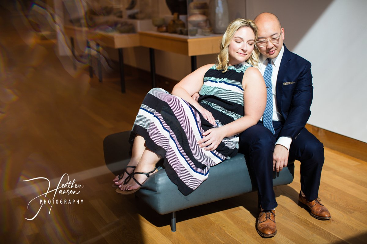 A couple sits close together on a small sofa in a museum gallery, the woman leaning her head on her partner's shoulder as he rests his hand on hers, both sharing a calm, tender moment.