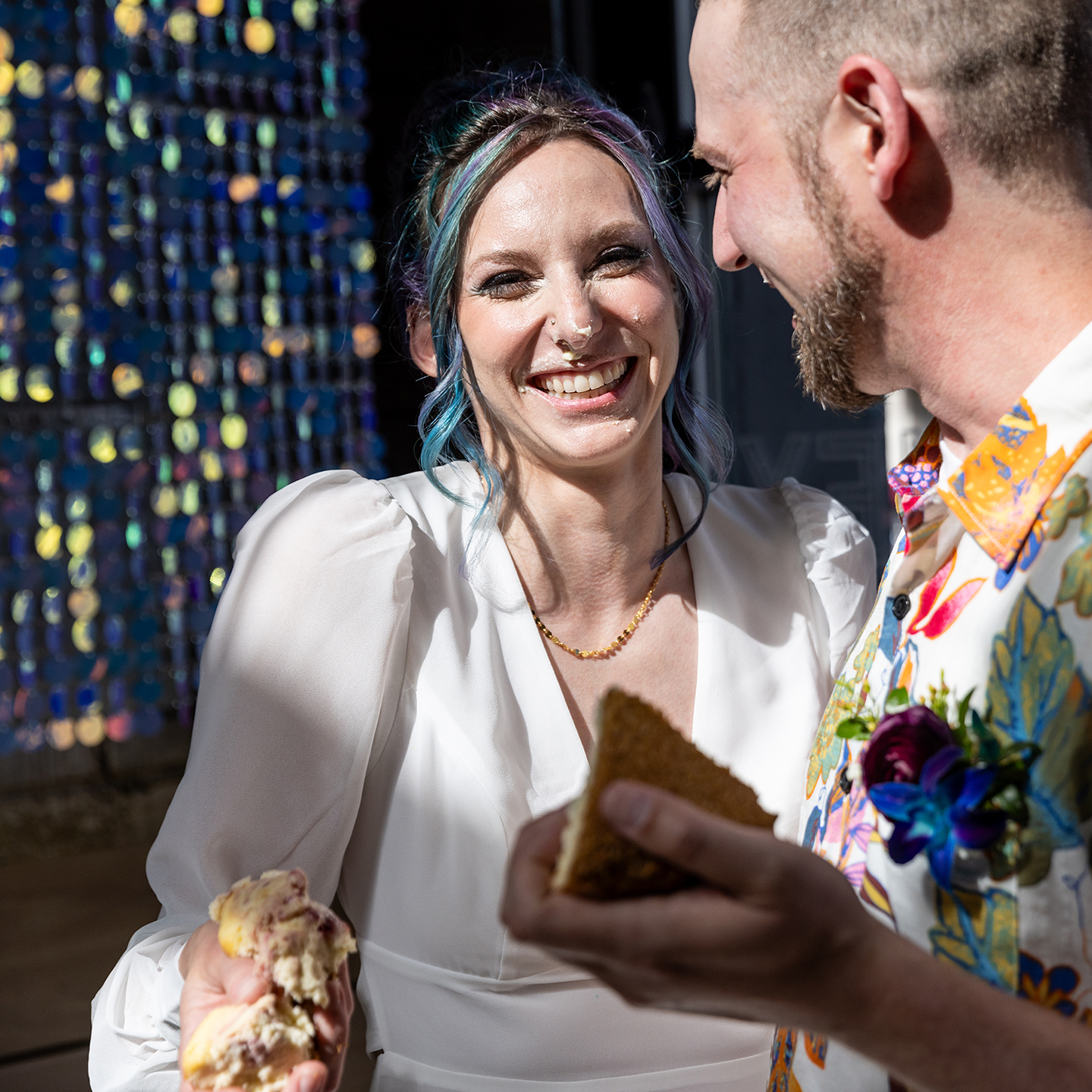 A bride with blue and purple hair smiles at camera with cheesecake on her face and in her clenched hand, her groom stands closely while looking at her.