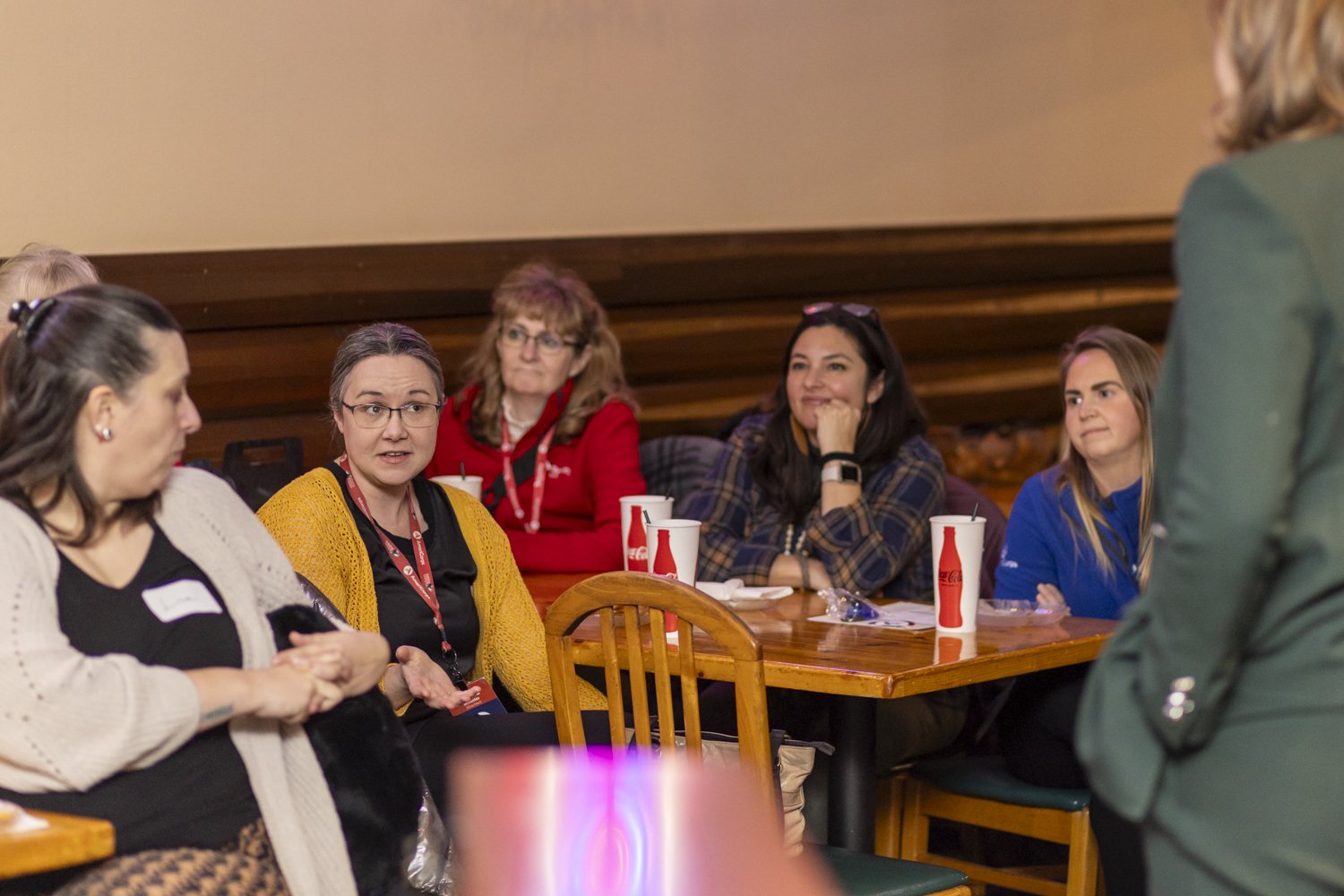 A group of women sitting at a table in a casual setting, attentively listening to someone off-camera. They have drinks in cups on the table.