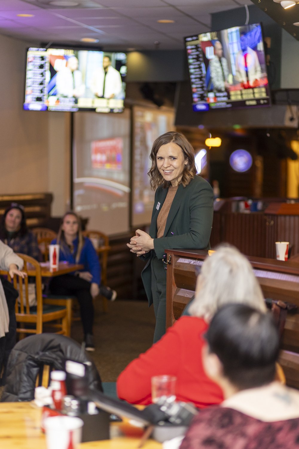A woman speaking at a casual event in a bar or restaurant, with attendees seated around tables listening and multiple TV screens displaying sports in the background.