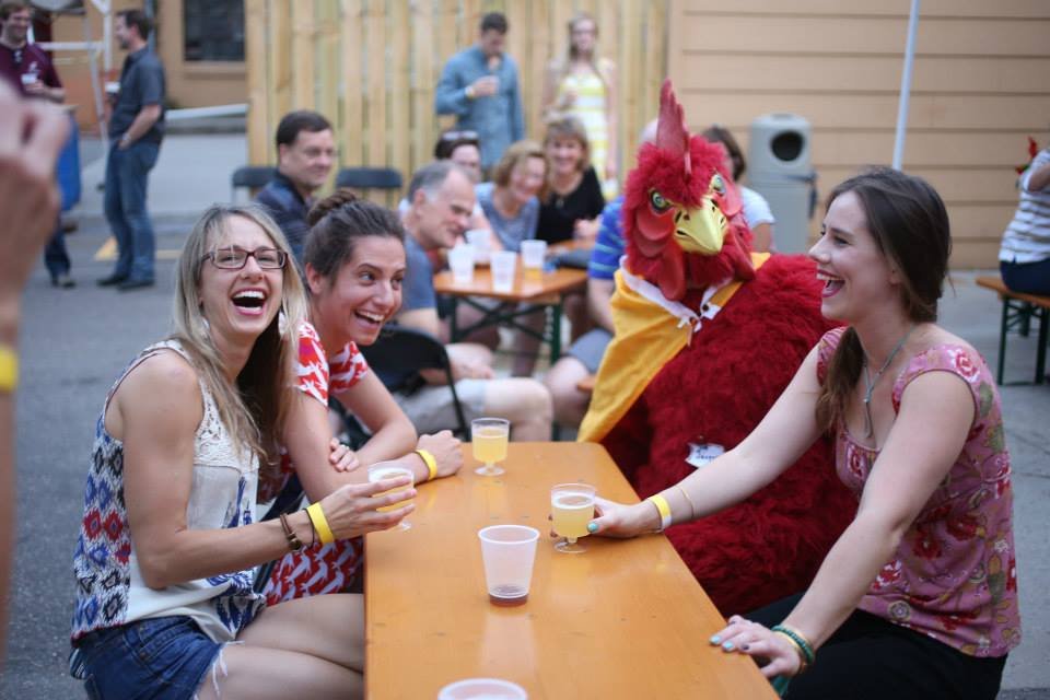 Group of people, including someone in a rooster costume, laughing and having drinks at an outdoor table.