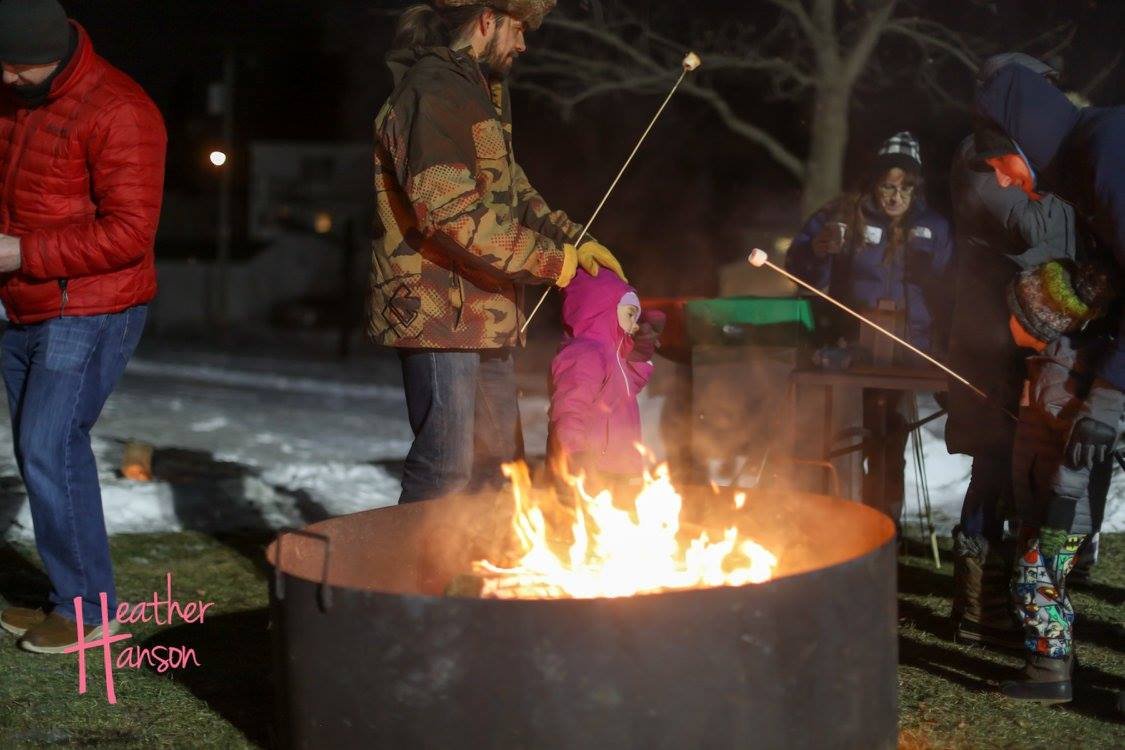 Group of people roasting marshmallows over an outdoor fire pit at night, with snowy background.