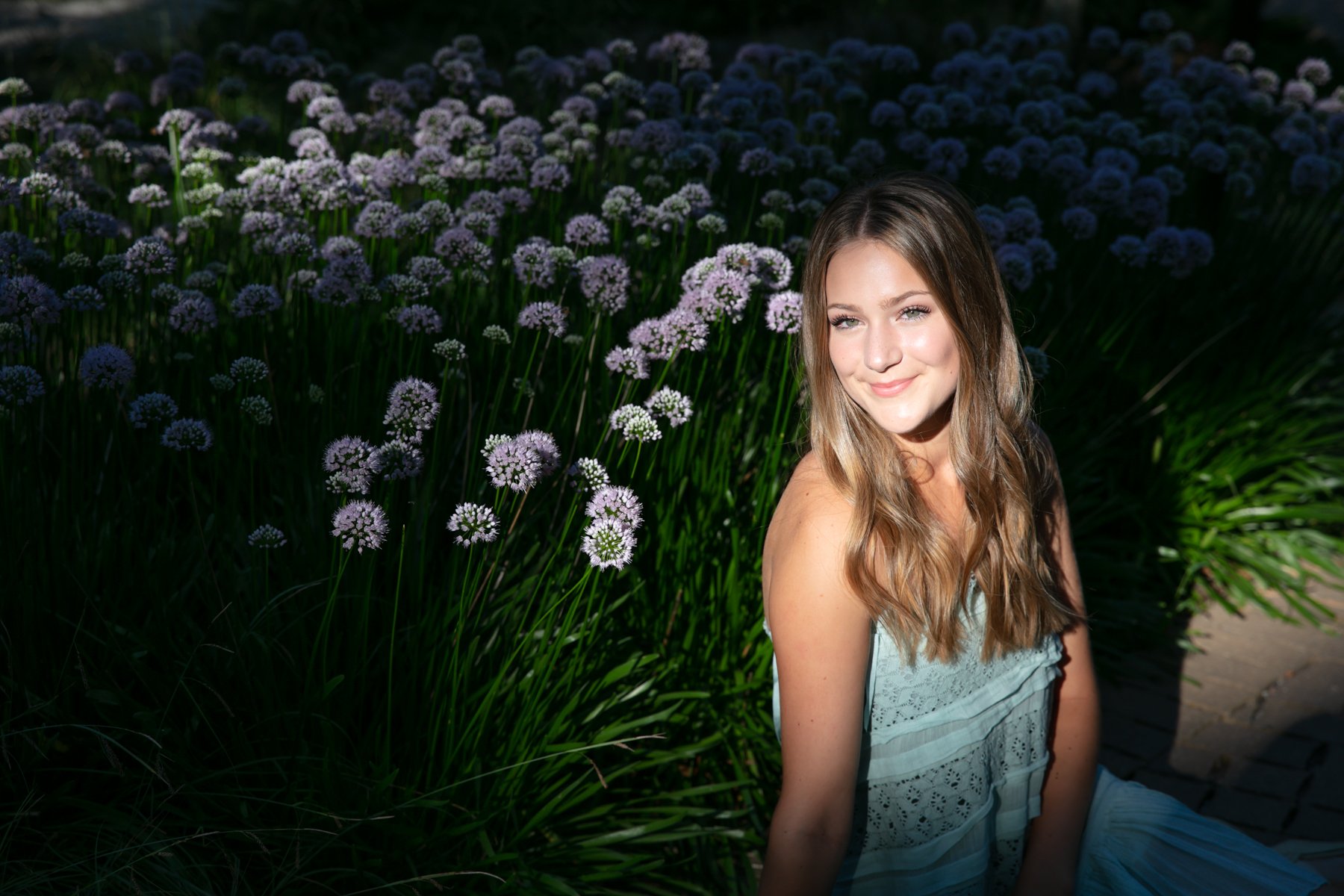 Young woman in a light dress sitting among purple flowers in a garden.