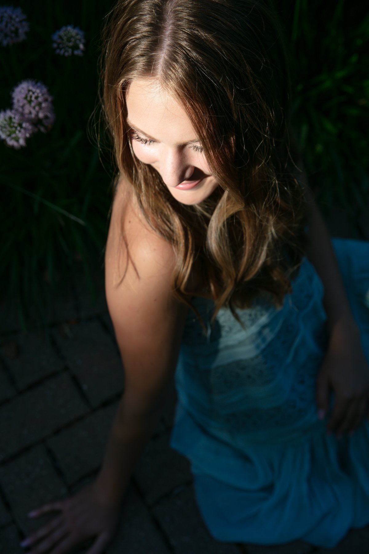 Woman in blue dress sitting on brick pavement with flowers nearby.