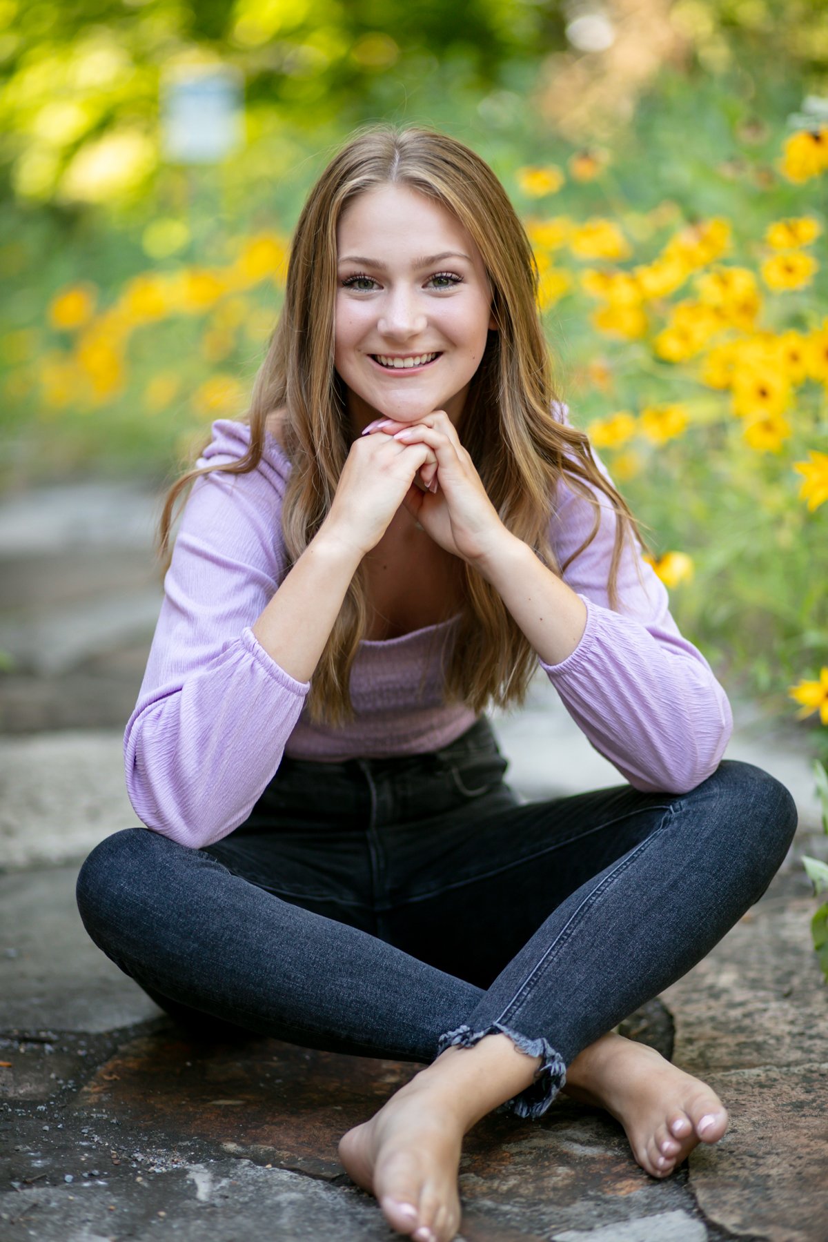 Smiling person with long hair sitting cross-legged on a stone path surrounded by yellow flowers.