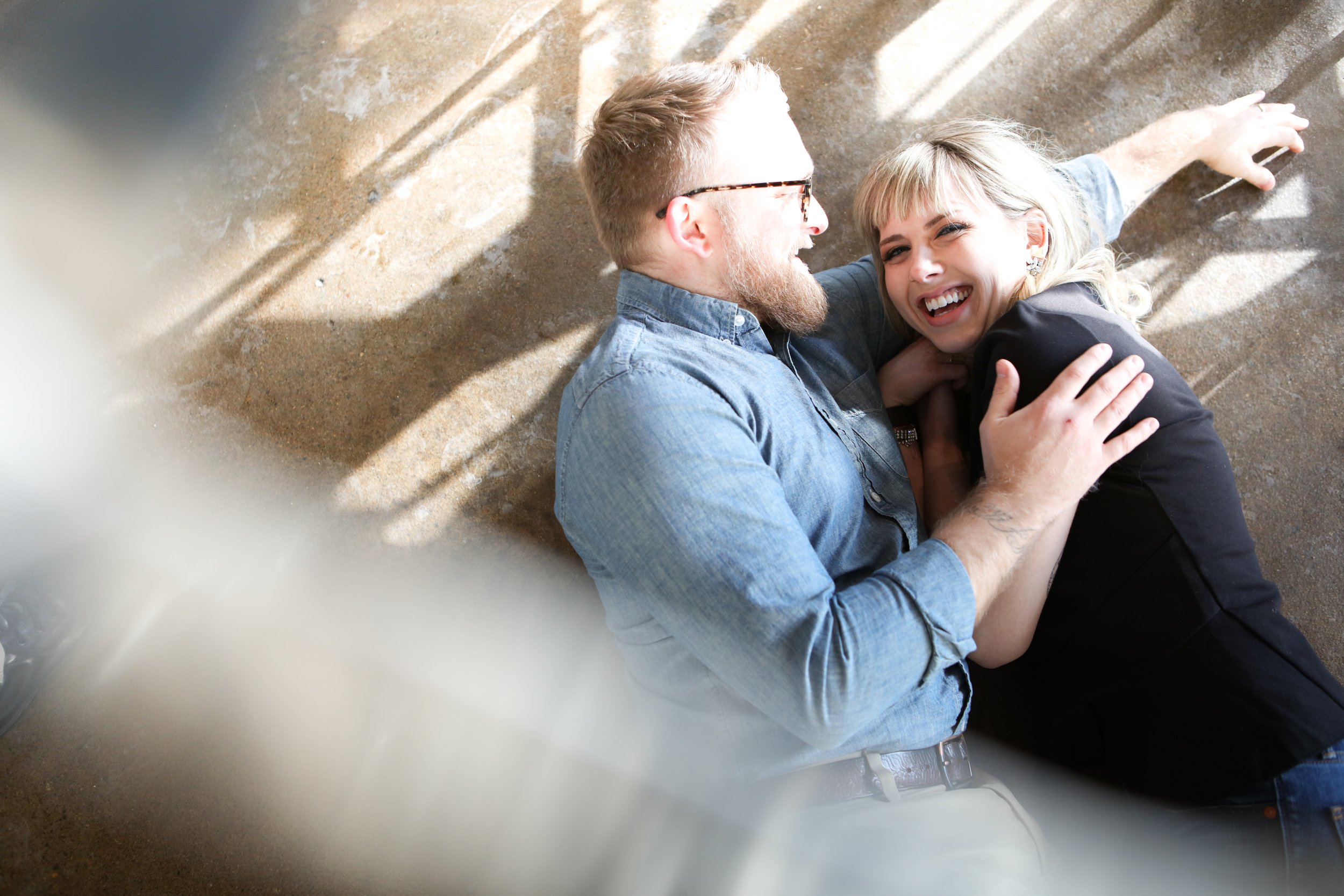 Man and woman lying on the floor, smiling and hugging each other, with sunlight casting shadows.