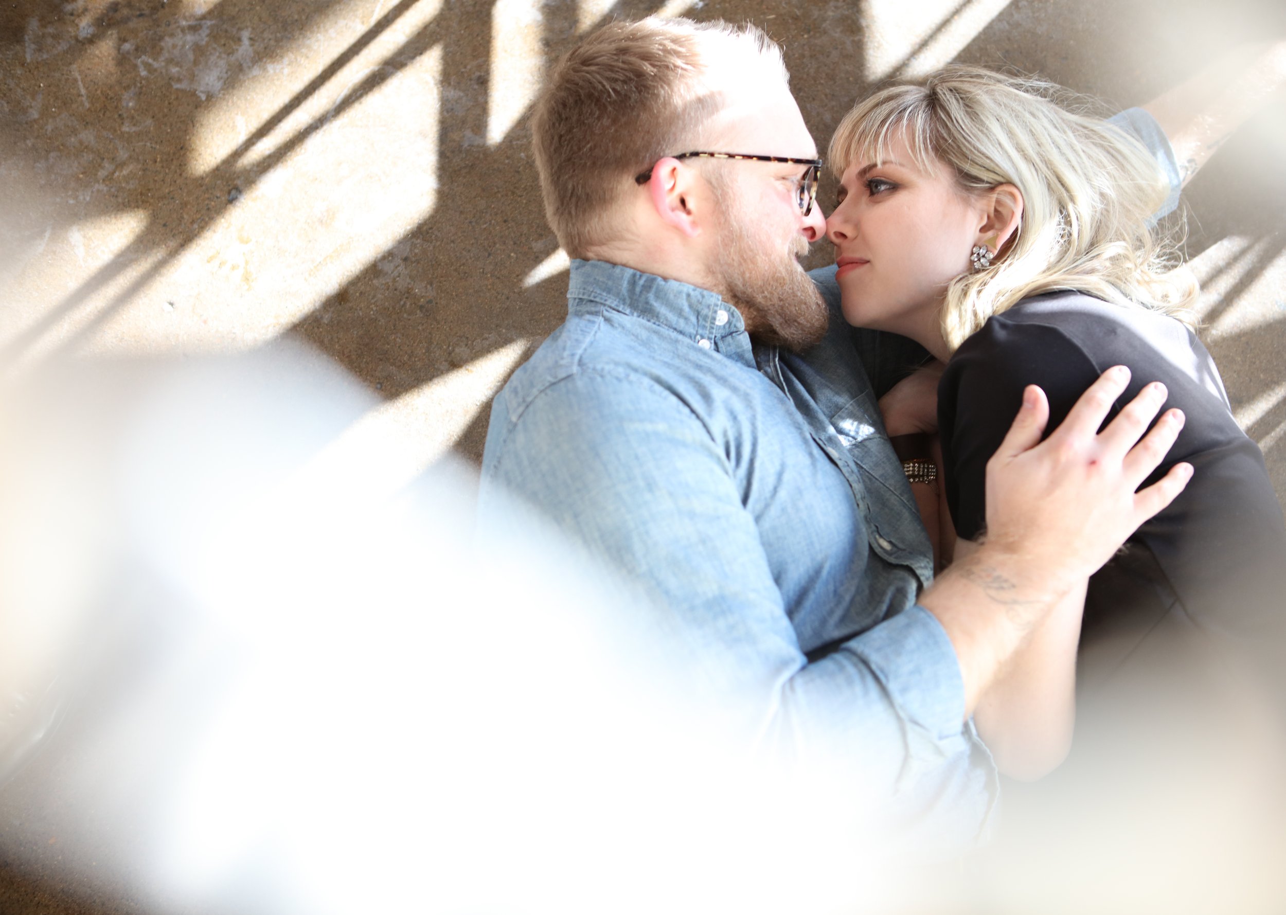 A couple lying on the floor, embracing and looking at each other affectionately, with sunlight casting patterns around them.