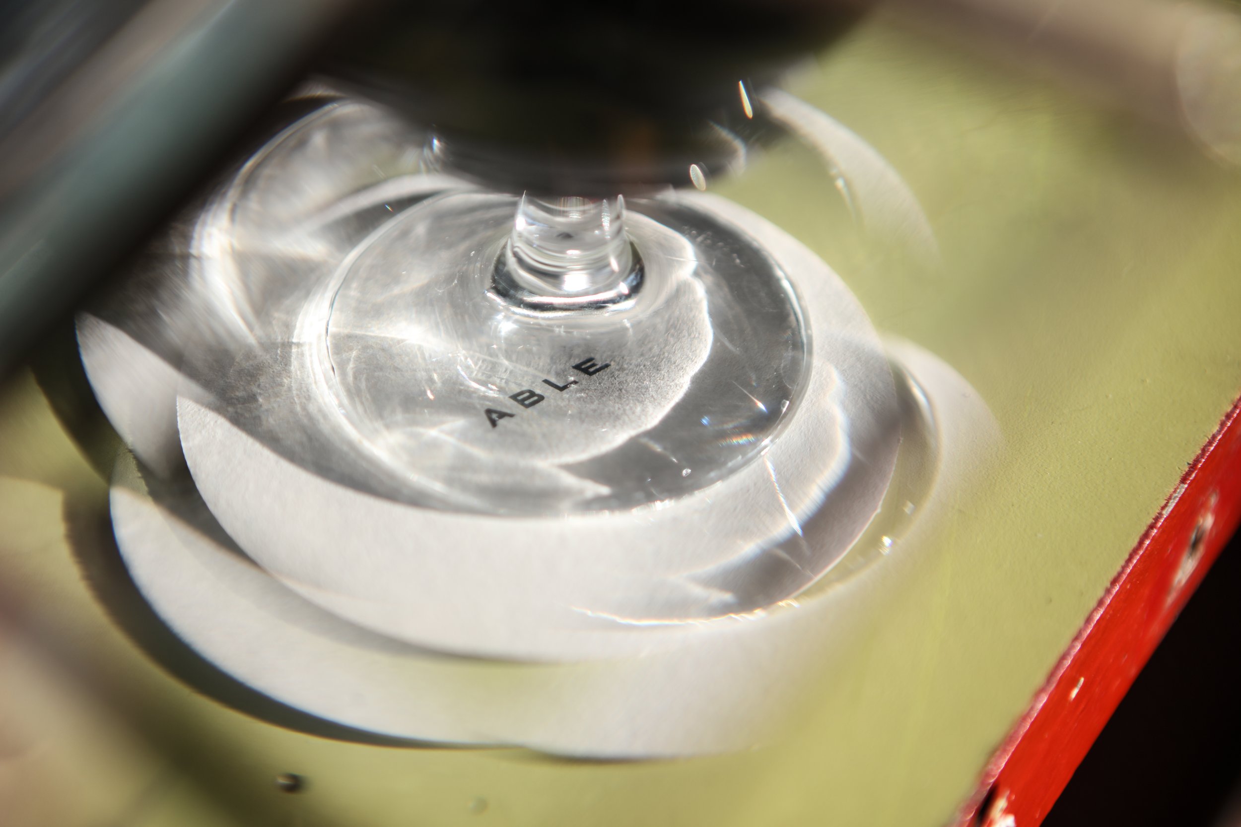 Close-up of a glass coaster with the word 'ABLE' printed on it, resting on a reflective surface.