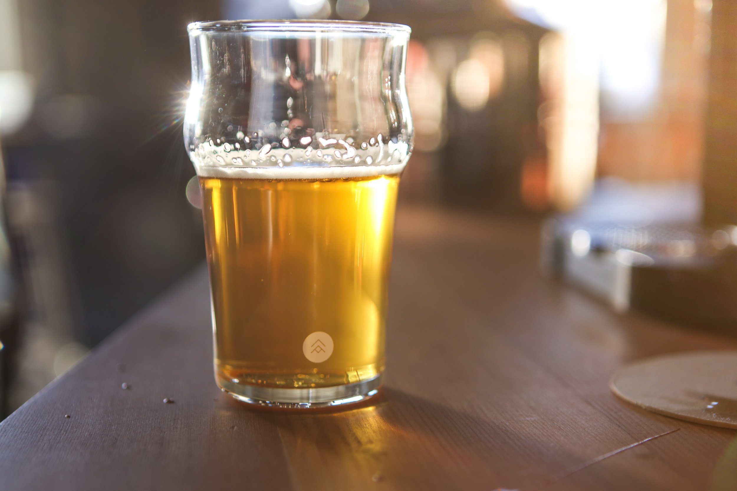 A pint glass of amber beer on a wooden table with sunlight reflecting off the glass.