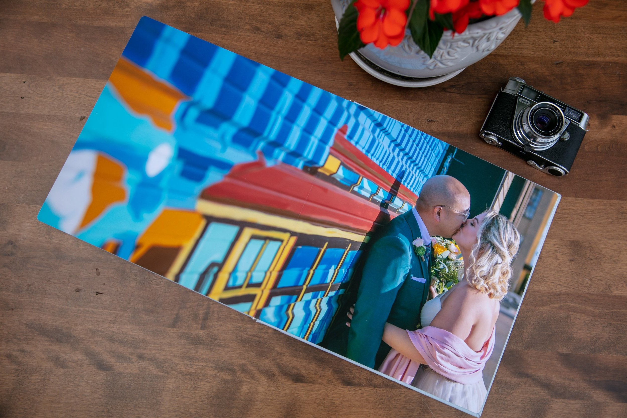 Open photo book with a picture of a couple kissing, next to a film camera and potted flowers on a wooden table.