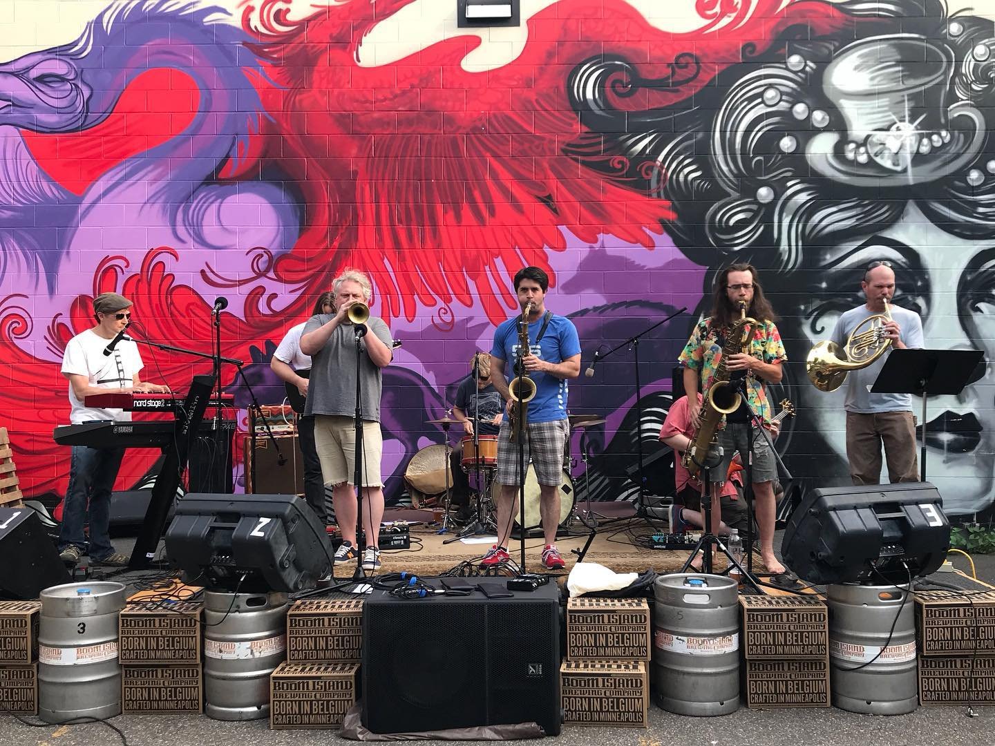 A band performing outdoors in front of a colorful mural, featuring musicians playing instruments such as keyboards, trumpet, saxophone, and French horn. The stage is decorated with beer kegs and boxes.
