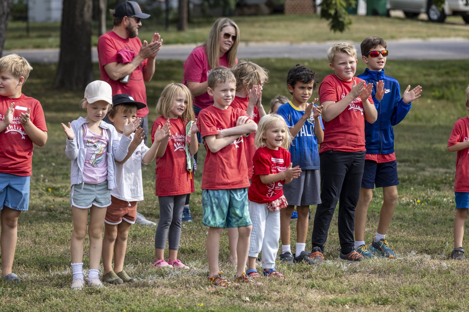 Group of children and adults clapping outdoors on a grassy field, some wearing red shirts.