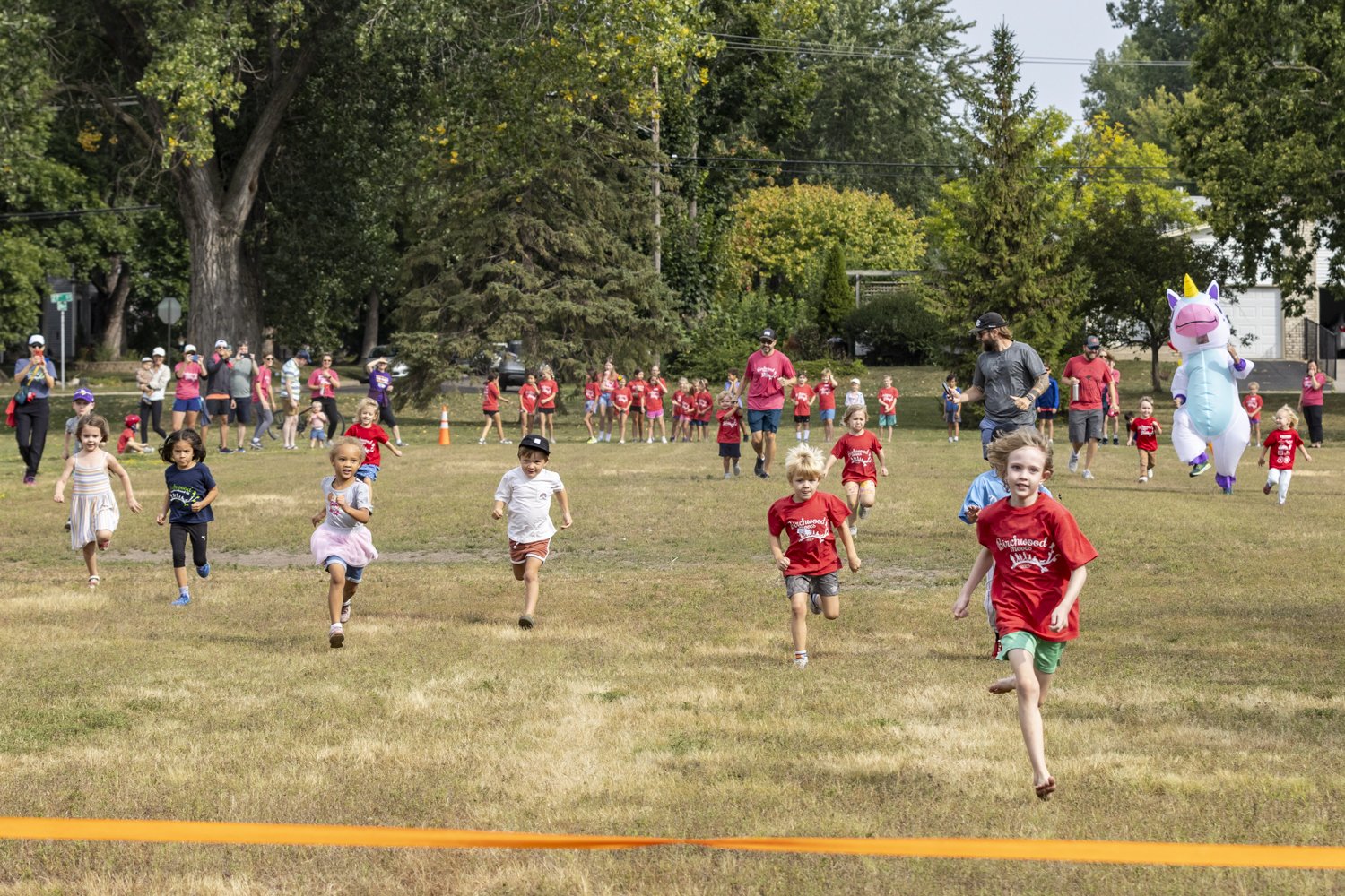 Children running race on a grassy field with spectators and a person in an inflatable unicorn costume in the background.