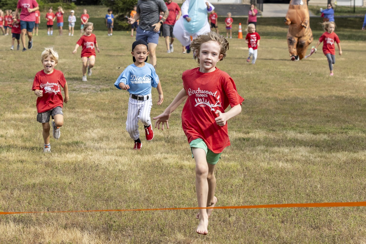 Children running a race on grass, wearing colorful shirts, including red and blue. Background features more kids and playful costumes.