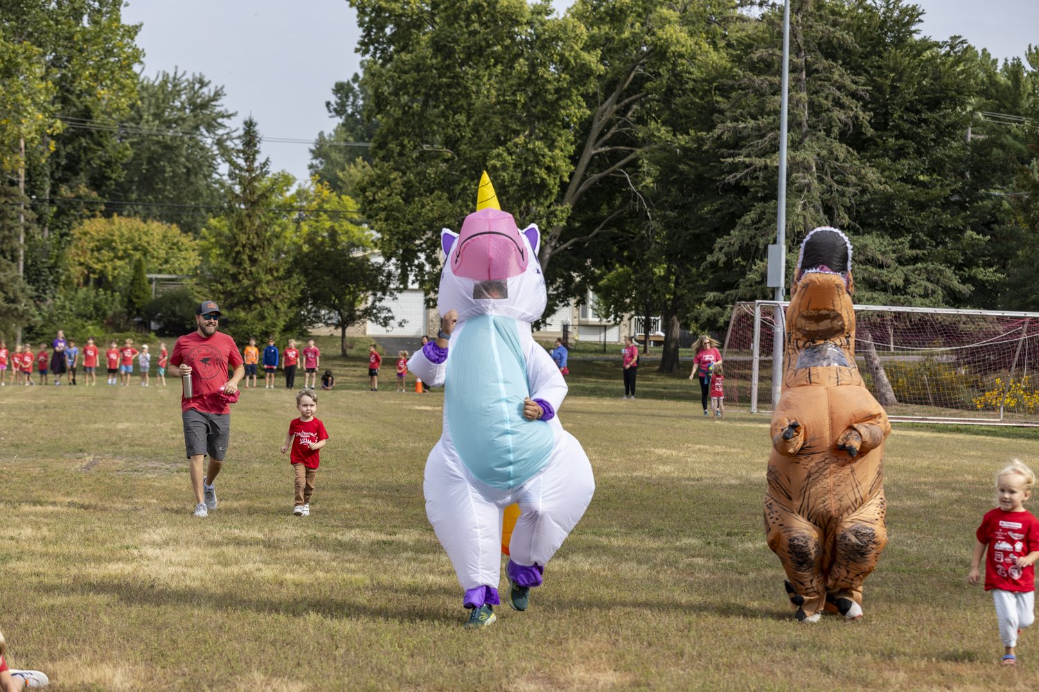 People in dinosaur and unicorn costumes running at an outdoor event alongside children in red shirts. Background includes spectators and soccer goalposts.