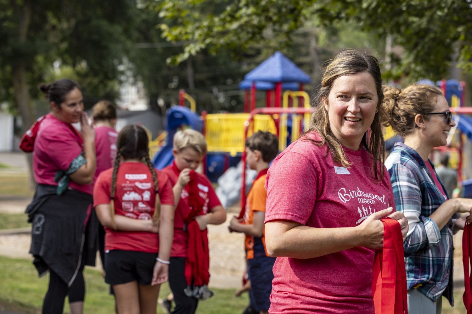 Group of adults and children in a playground, people wearing red and blue shirts, with play structures in the background.