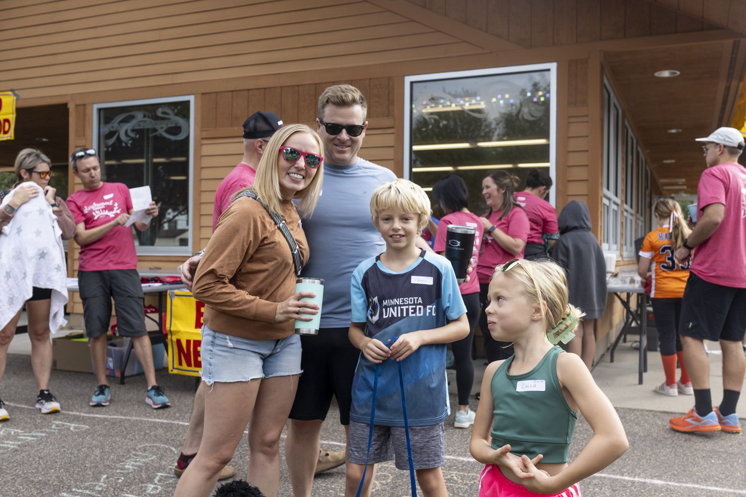 Outdoor family gathering with people socializing and children playing, participants wearing casual attire, snacks and drinks on tables, summer vibe.