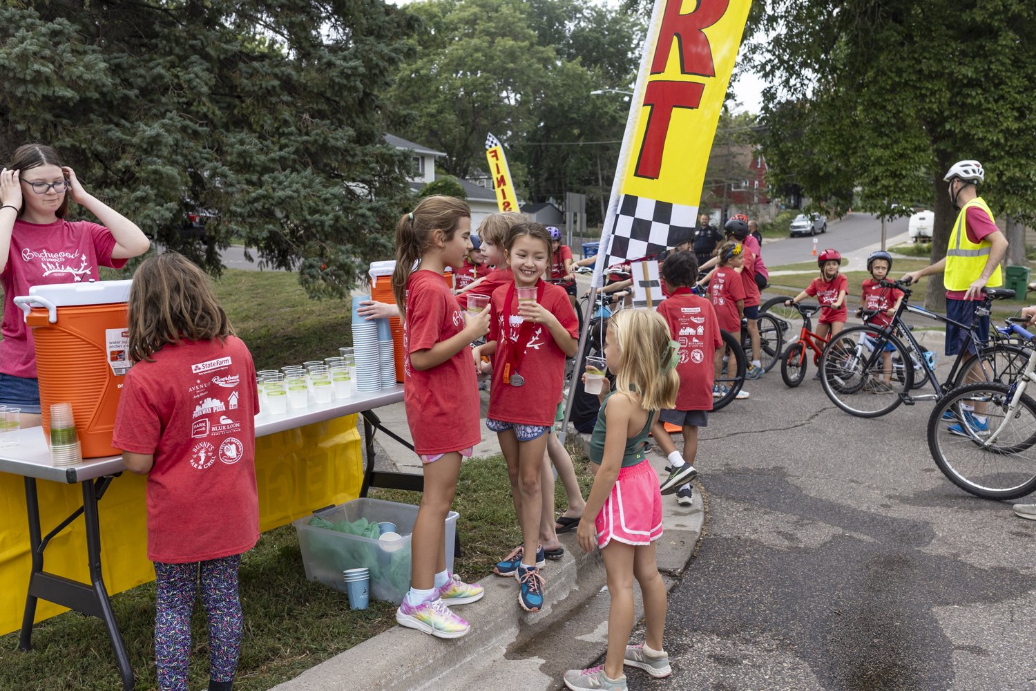 Children gathered at a beverage station at a fun run event, wearing red shirts and helmets, with bicycles nearby. A start flag is visible.