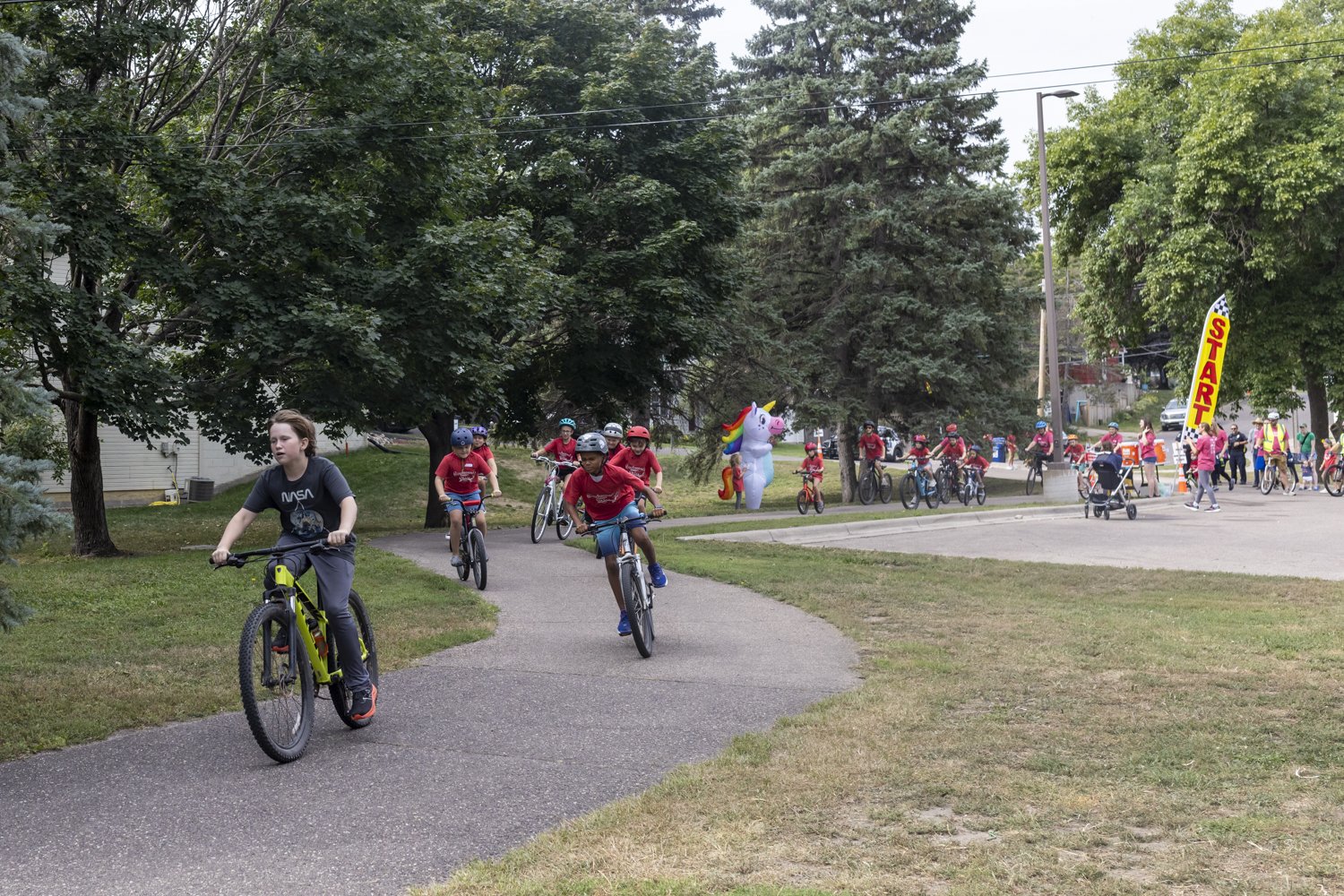 A group of people participating in a biking event at a park, with a giant inflatable unicorn and a "START" banner. Most participants are wearing helmets and red shirts, cycling on a path surrounded by trees.
