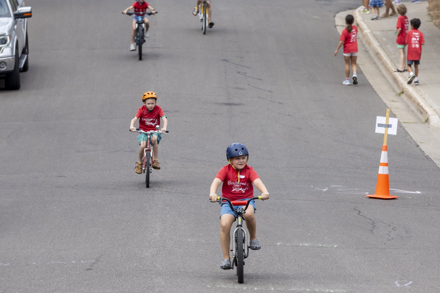 Children riding bicycles on a street, wearing helmets, with traffic cones and others walking on the sidewalk.