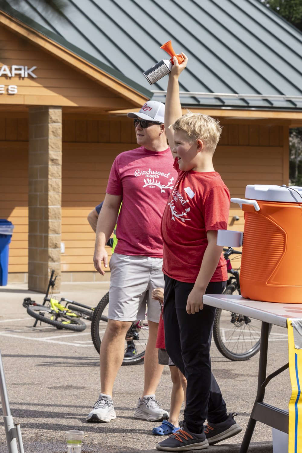 Boy holding an air horn next to a man in matching red shirts, standing near a table with an orange cooler. They're at an outdoor event with bicycles in the background.