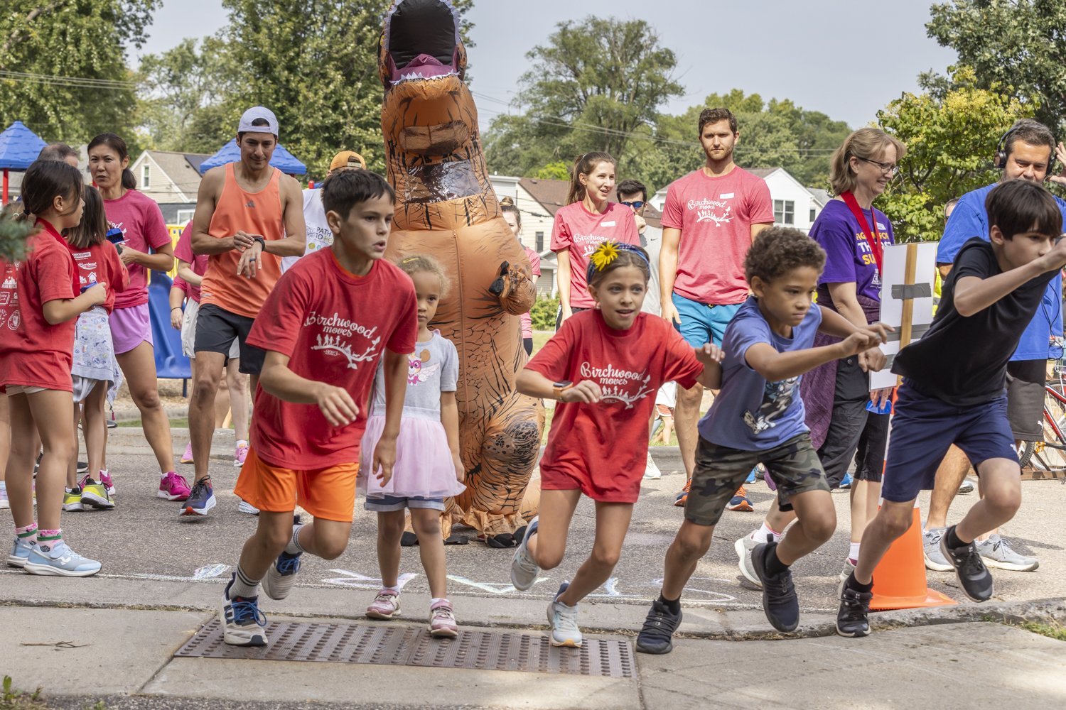 Children starting a race at a community event with spectators and a person in a dinosaur costume.