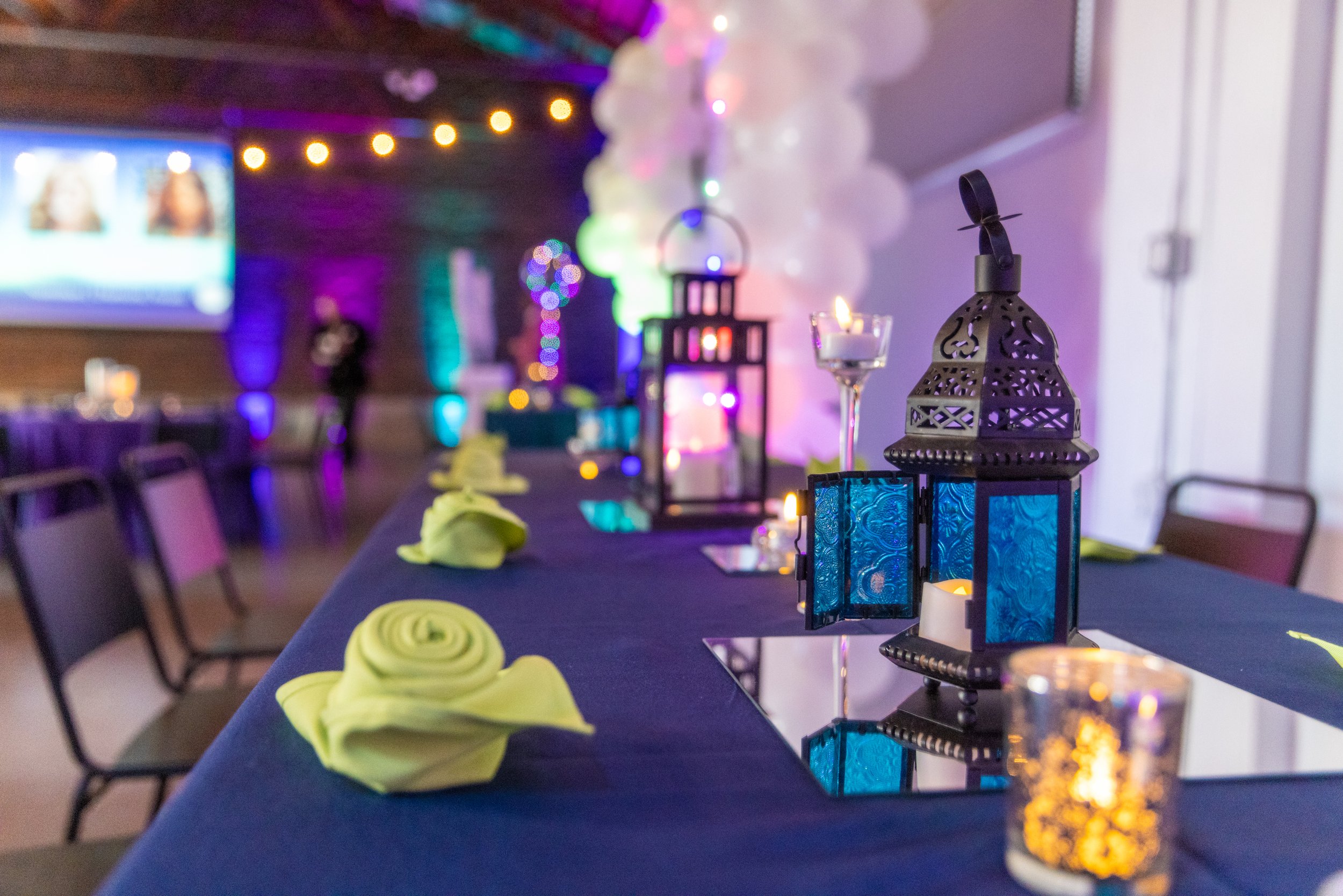 Decorative table setting at an event with blue tablecloth, green rolled napkins, lanterns, and candles. Background features colorful lighting and balloons.