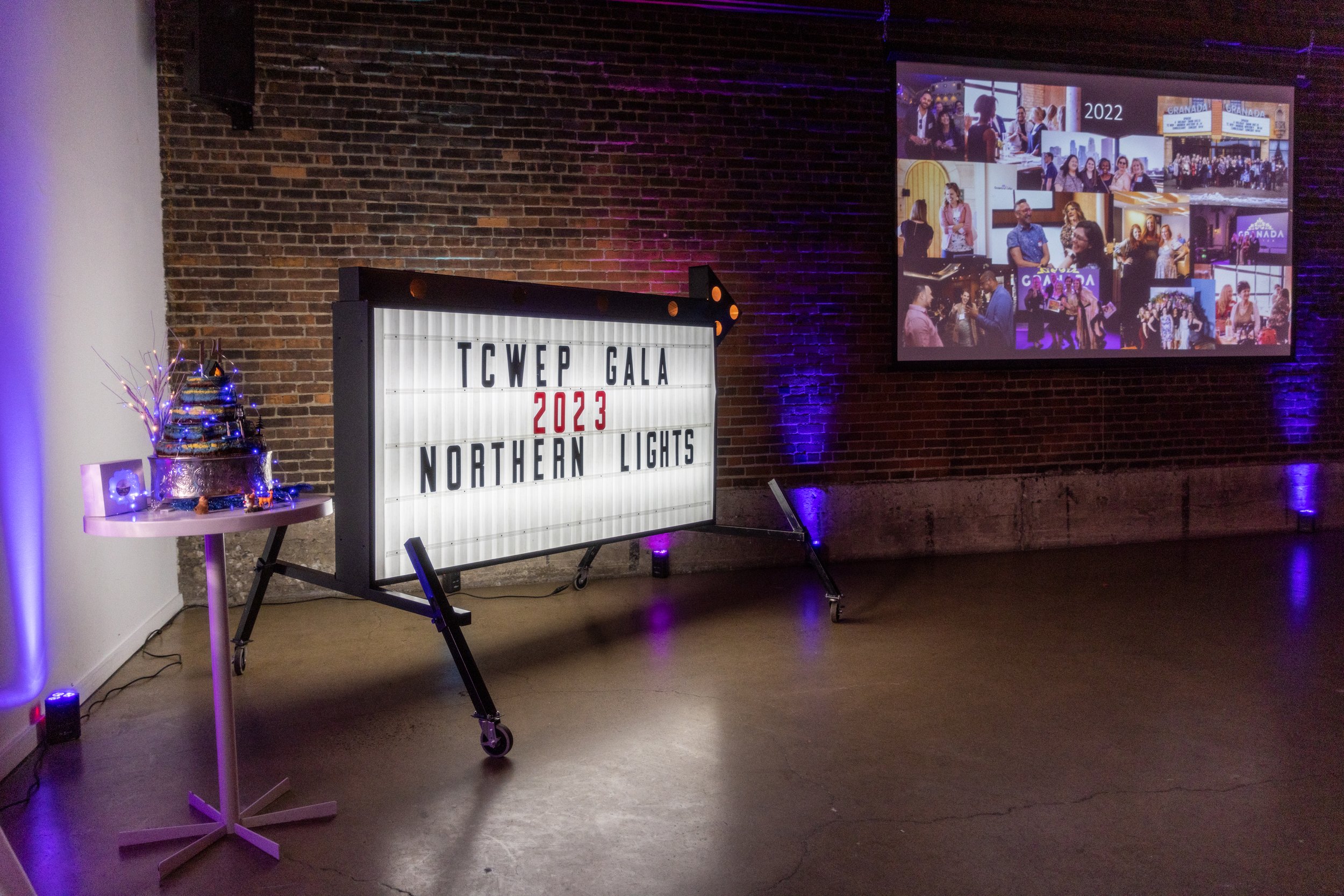 Large illuminated sign displaying "TCWEP Gala 2023 Northern Lights" with a decorated cake on a table, and a photo collage projected on the wall in a dimly lit brick-walled room.