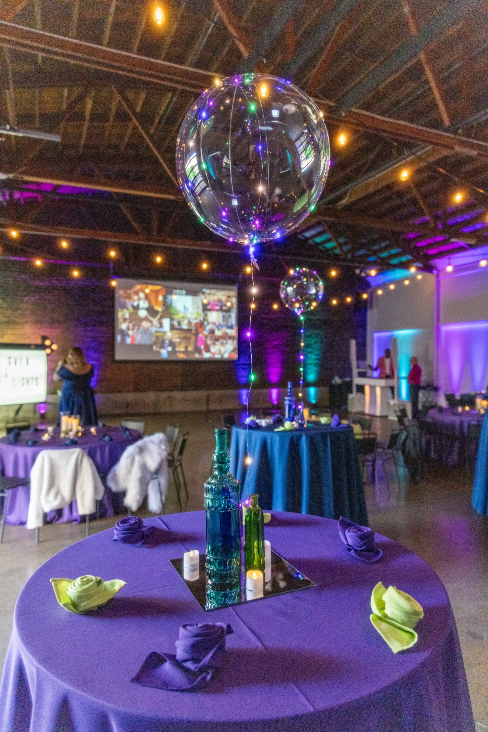 Event venue with tables covered in purple tablecloths, decorated with glass bottles and folded napkins; large inflatable balloons with lights; fairy lights overhead; projected images on a wall.