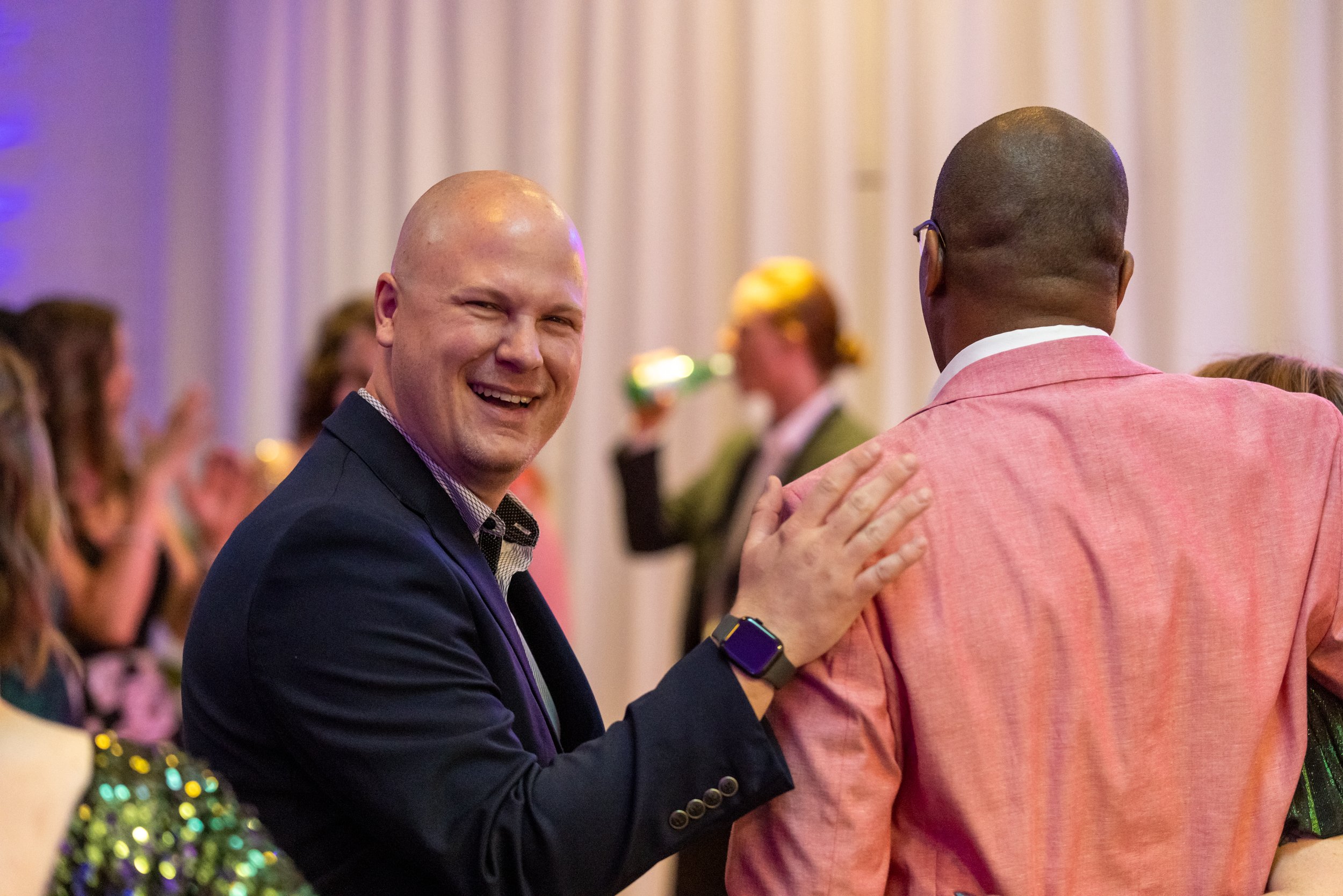 A smiling man at a social event with a crowd in the background.