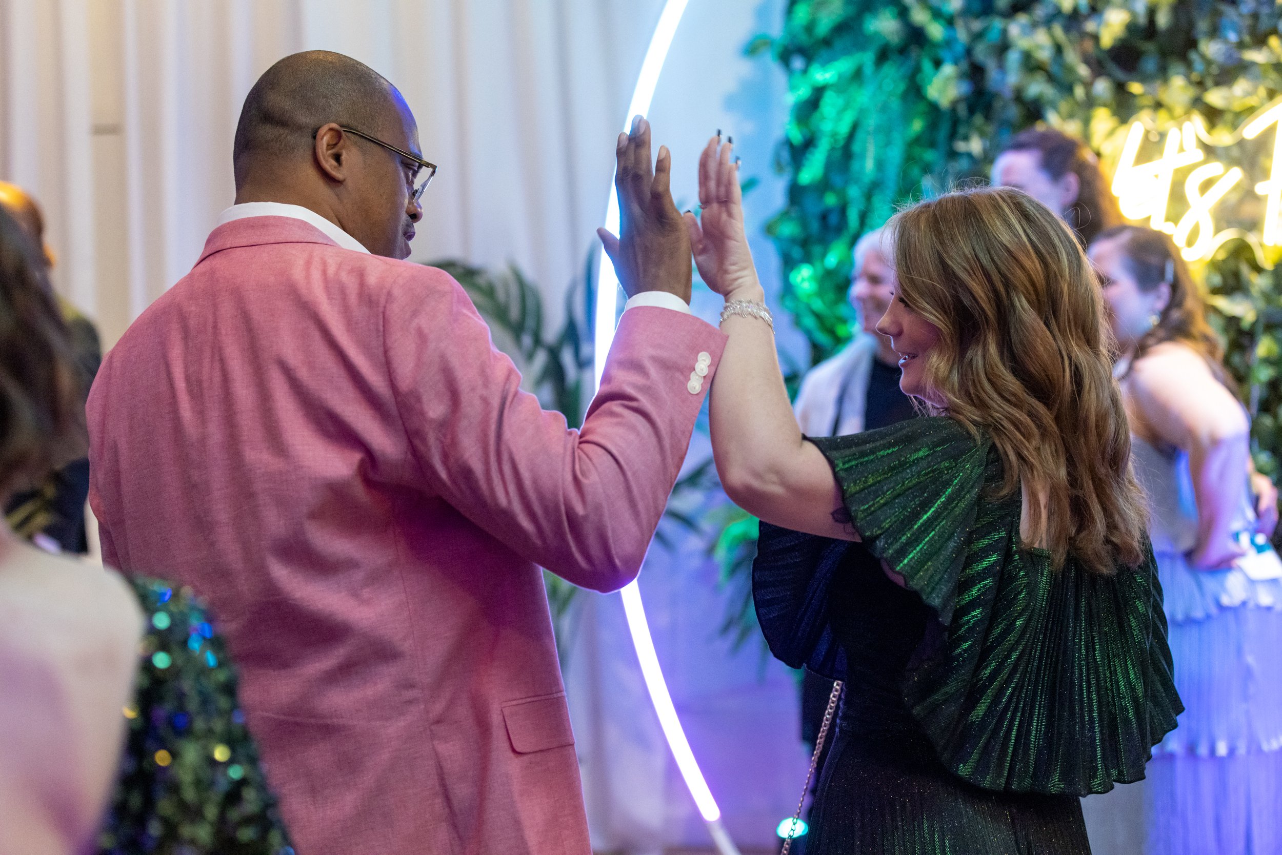 Two people high-fiving at an event, with colorful lighting and greenery in the background.