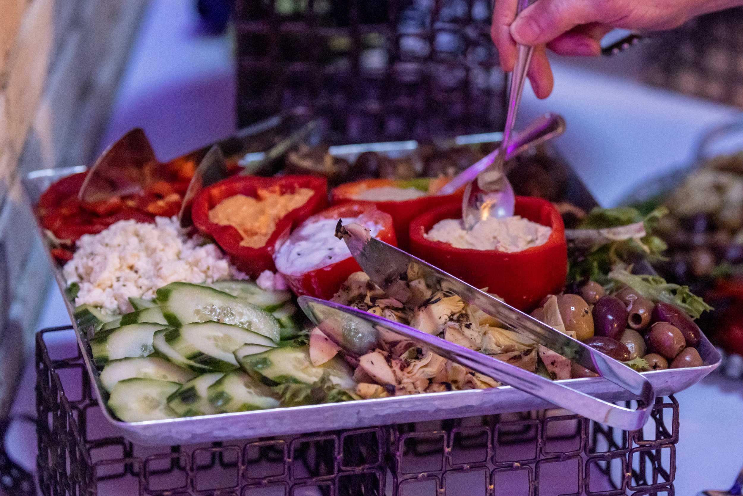 Assorted appetizers on a tray, including sliced cucumbers, stuffed red bell peppers, feta cheese, olives, and a hand holding a spoon.