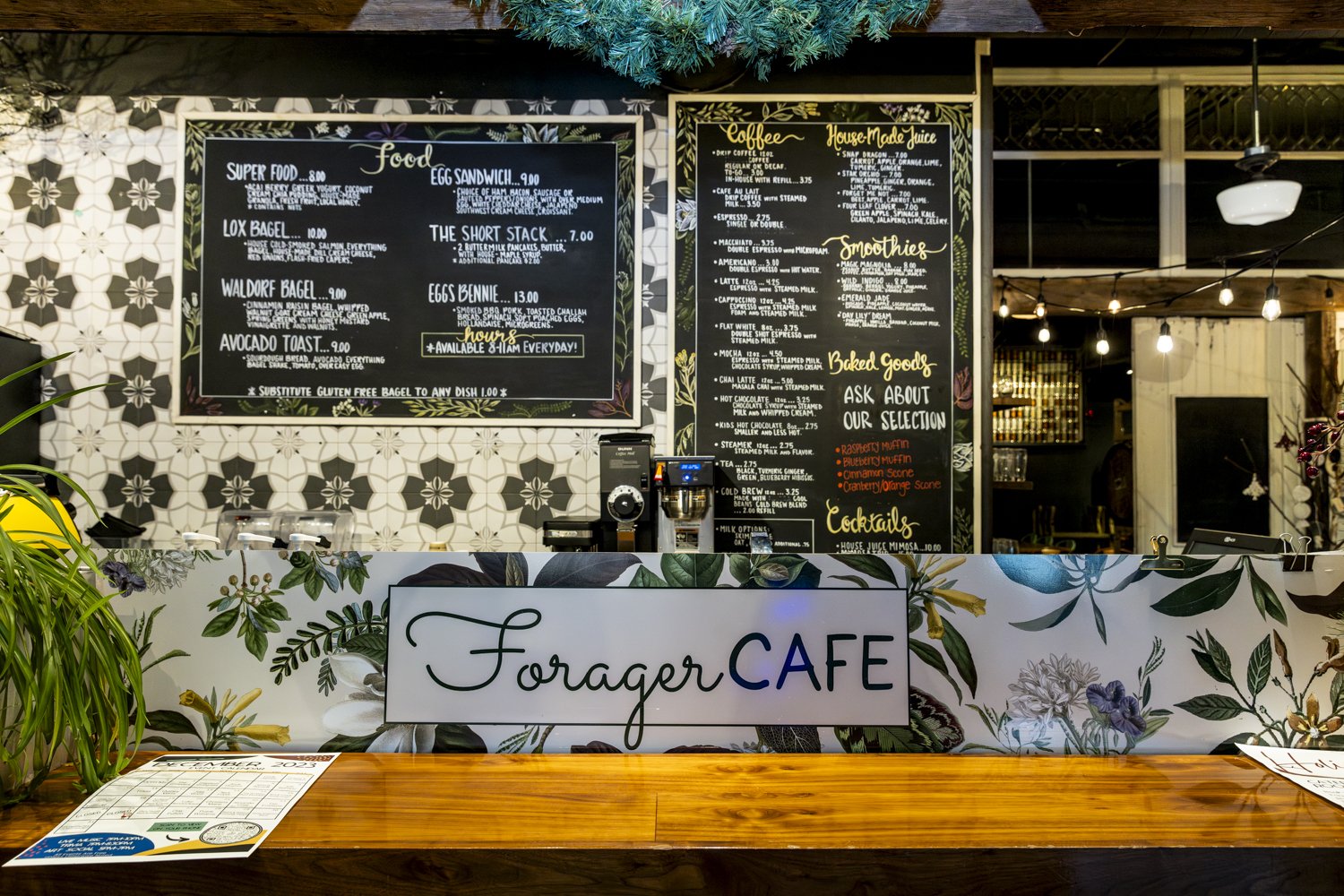 Forager Cafe counter with menu boards displaying food, coffee, smoothies, baked goods, and cocktails; decorative floral wallpaper and a wooden counter.