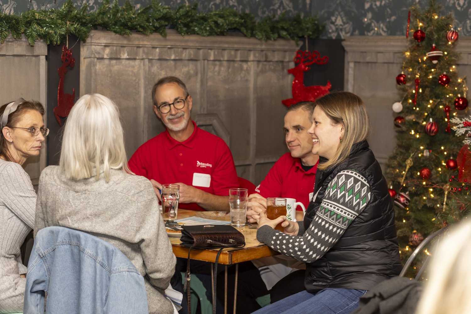 Group of people sitting at a table, talking and smiling, with Christmas decorations and a tree in the background.