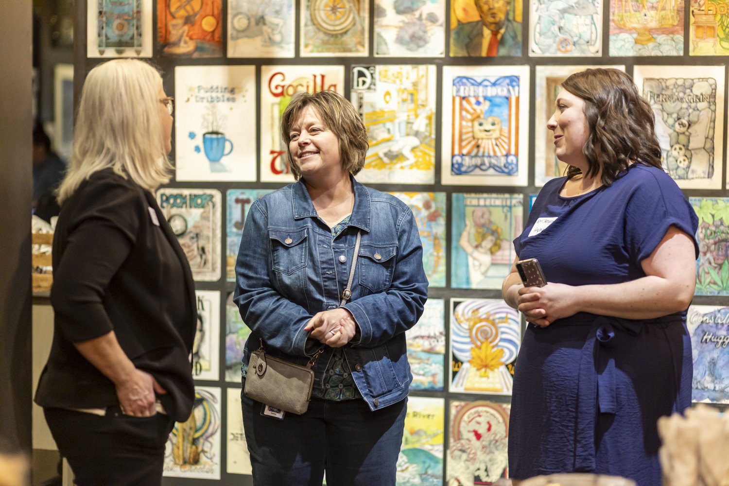 Three women conversing in front of an art display with various framed illustrations on the wall.