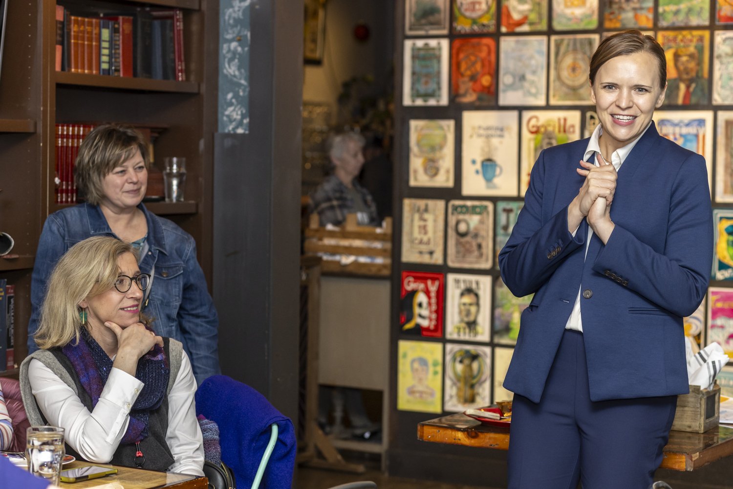 A woman in a blue suit smiles in a room with colorful artwork on the wall. Two other women are seated nearby, one wearing glasses and a scarf. Books are visible on a shelf.