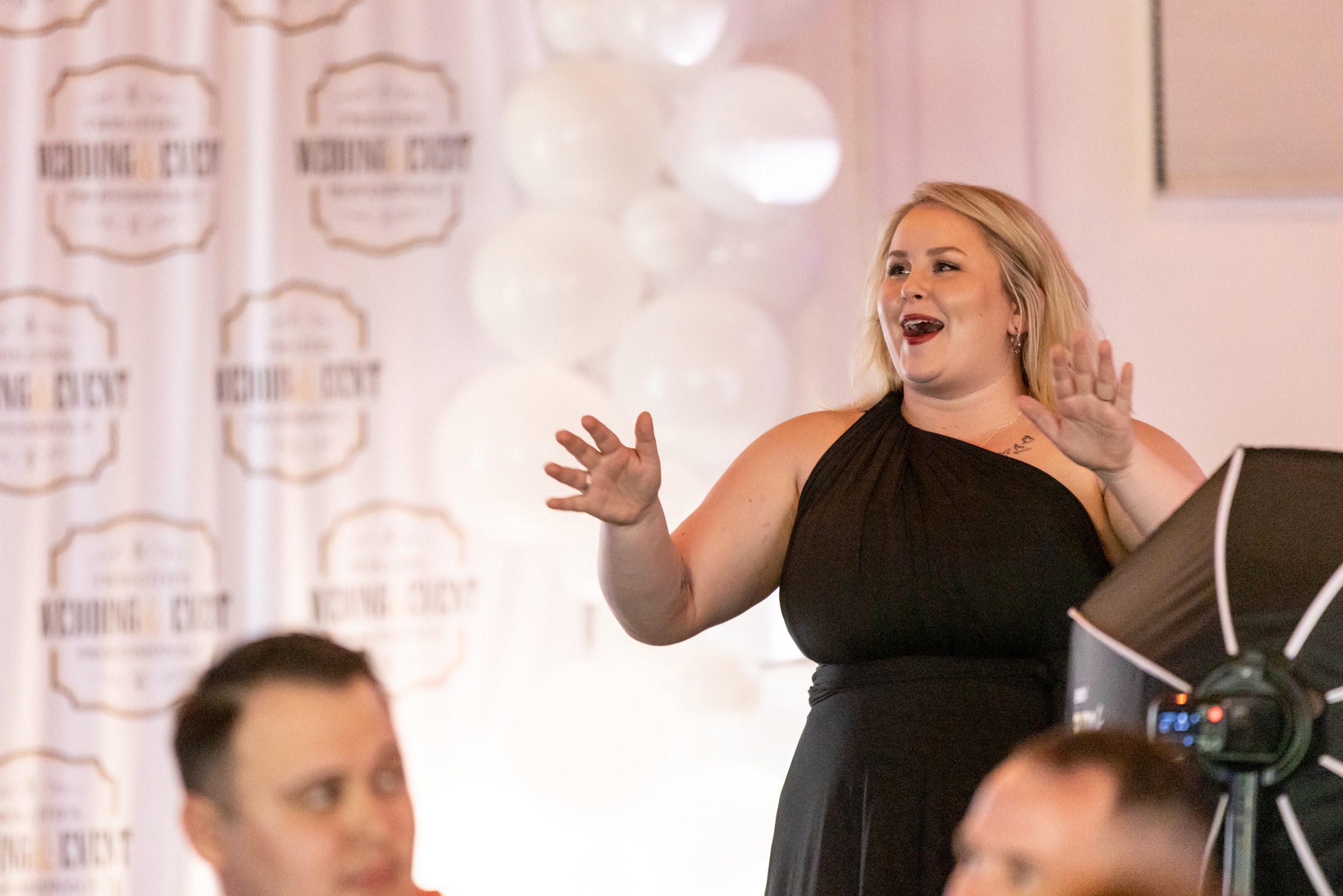 Woman in black dress speaking with excitement at event, with background decorations and a banner.