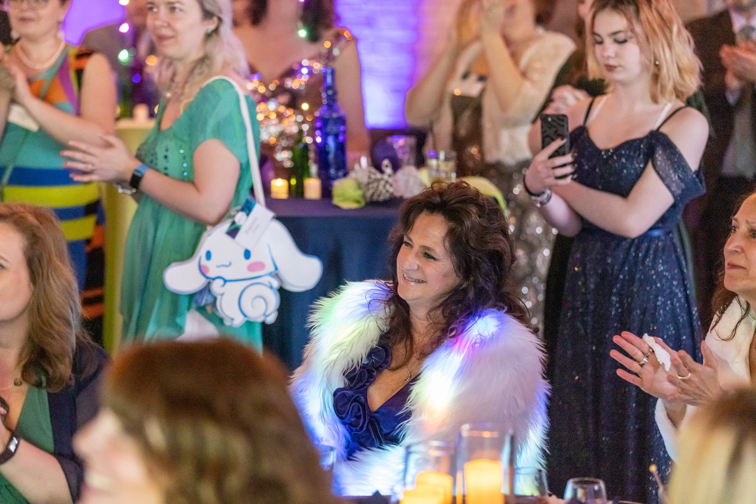 People at a festive event with colorful lighting, wearing elegant outfits; one person has a Cinnamoroll bag. They are clapping and taking photos with a phone.