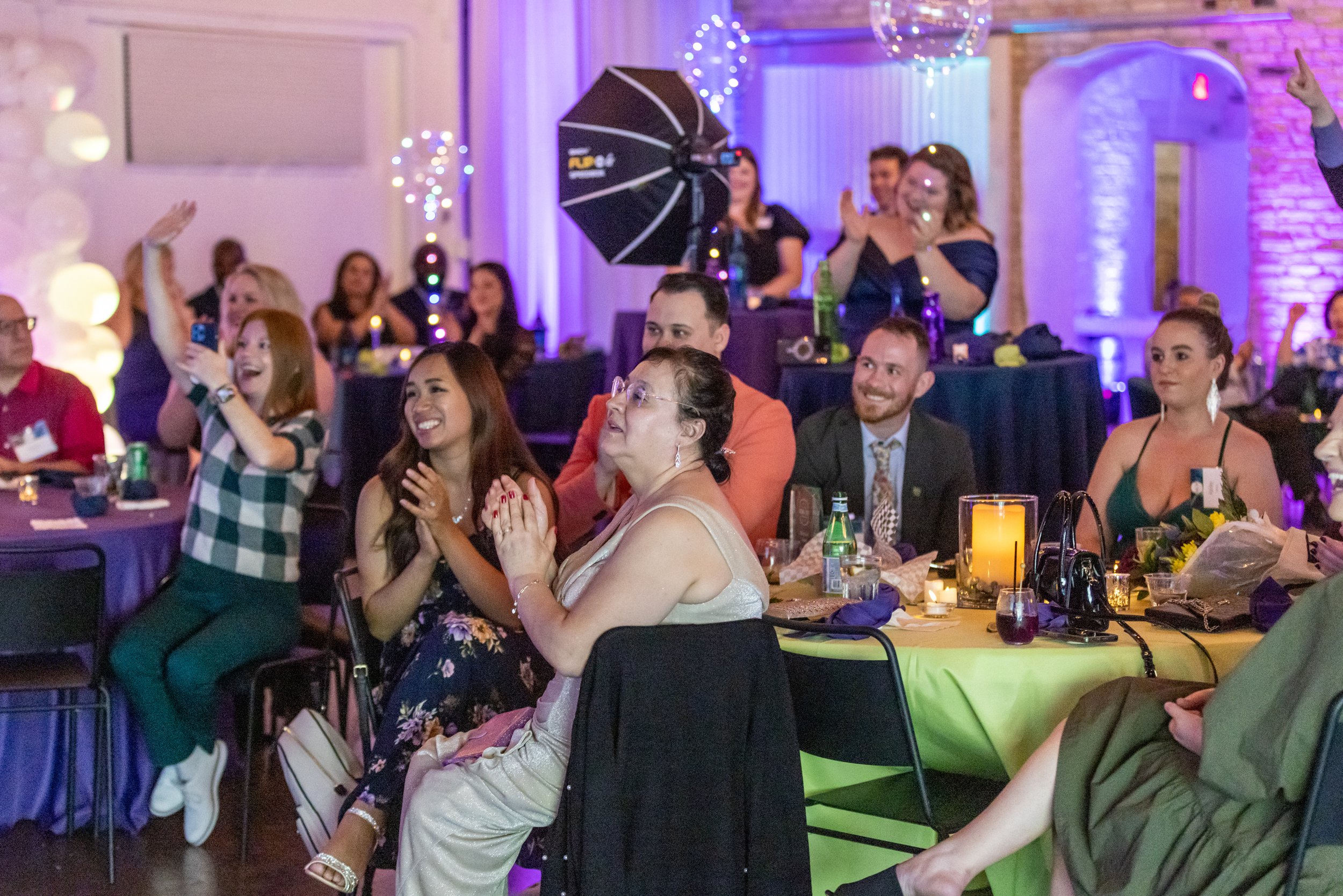 A group of people sitting at tables with drinks and decorations, clapping and smiling at an indoor event with purple lighting.