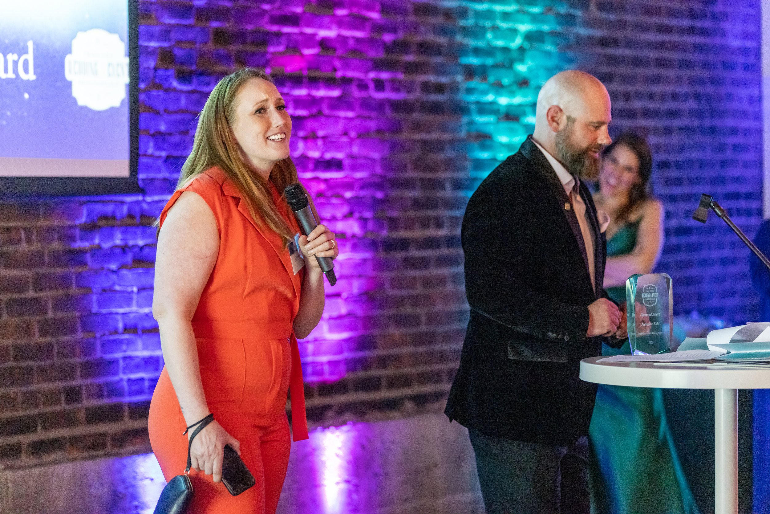 Two people speaking at an event with colorful lighting, one holding a microphone, and a woman in the background. An award is on the table.