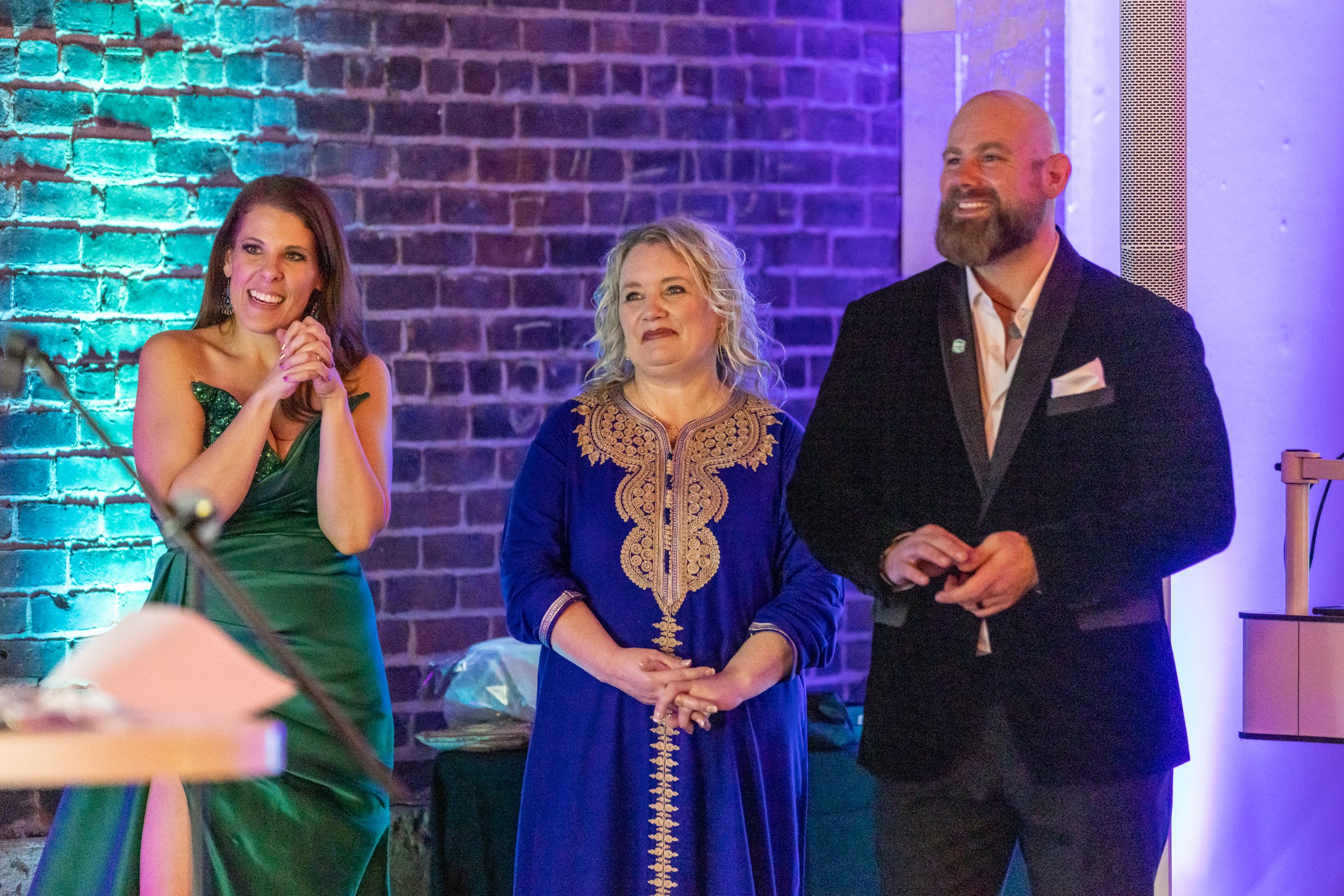 Three people dressed in formal attire at an event, standing against a brick wall with teal and purple lighting.