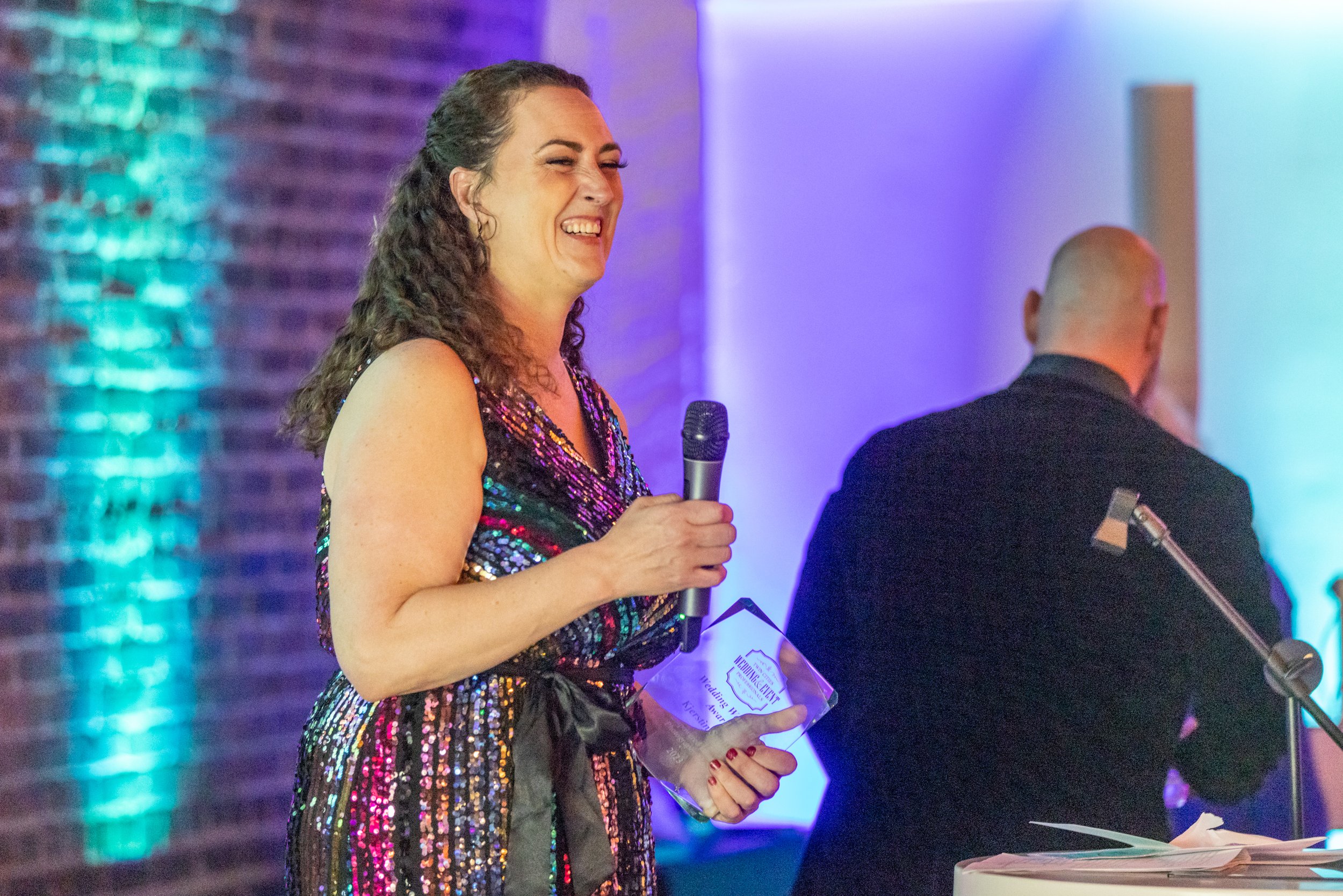 Woman in colorful sequin dress holding microphone and glass award, smiling at event