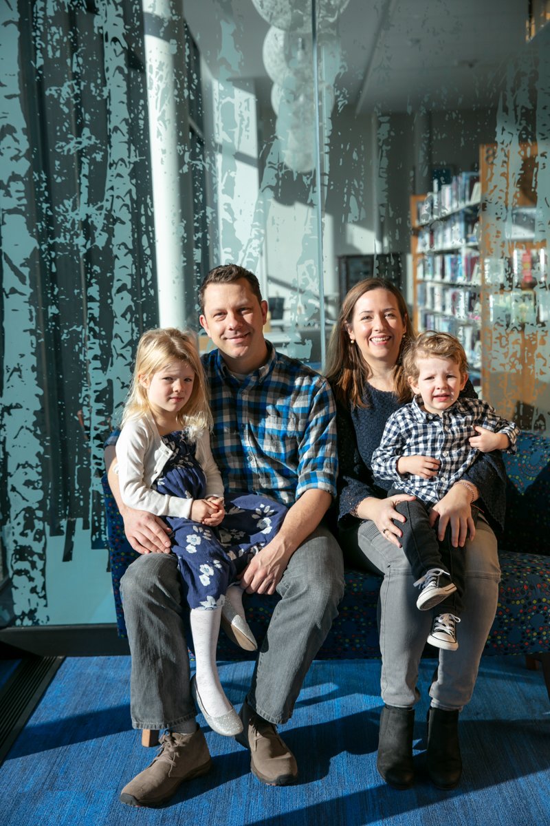A family of four sitting together indoors with abstract art and bookshelves in the background. The father and daughter wear plaid, while the mother and son are dressed in blue. They are all smiling.