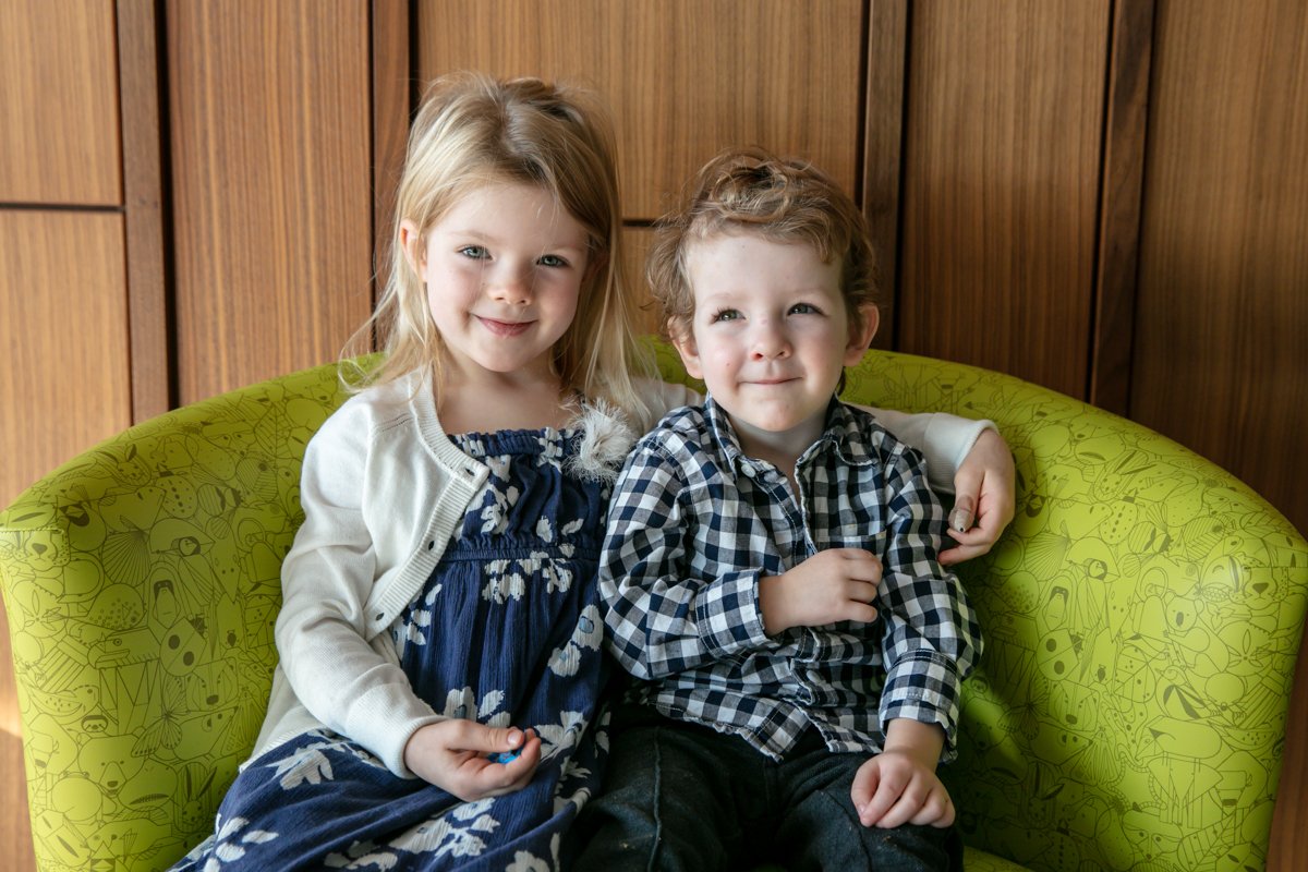 Two young children sitting on a green patterned chair, smiling at the camera. The child on the left is wearing a blue floral dress and white cardigan, and the child on the right is wearing a plaid shirt.