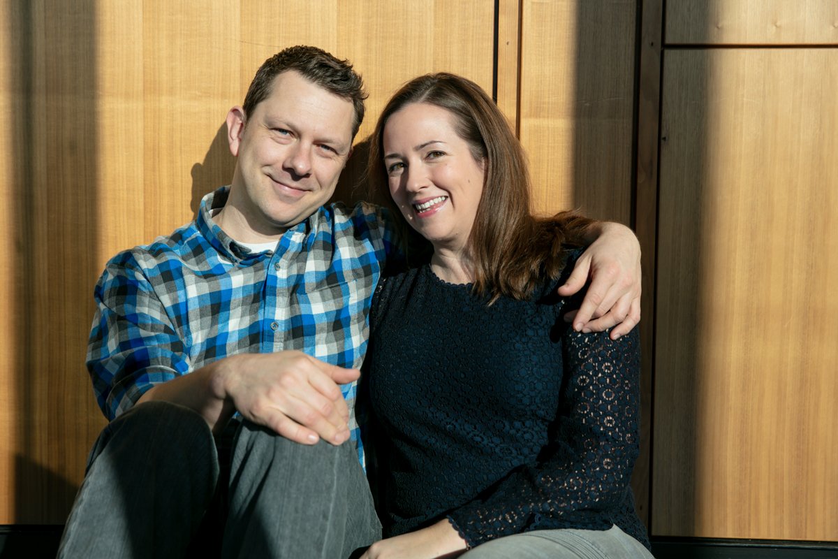 A man and woman sitting together, smiling, with the man wearing a blue plaid shirt and the woman in a dark lace top, against a wooden backdrop.