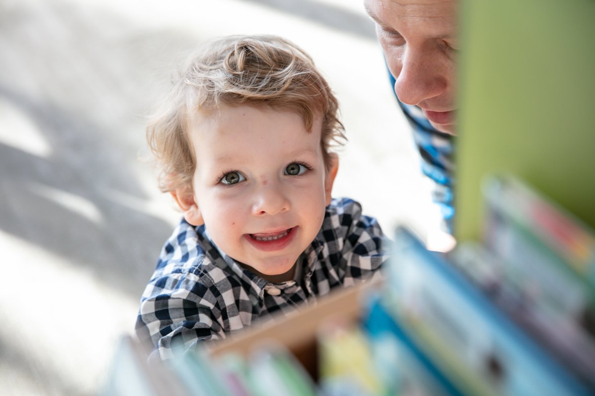 Young child smiling in a checkered shirt with an adult nearby, near a bookshelf.