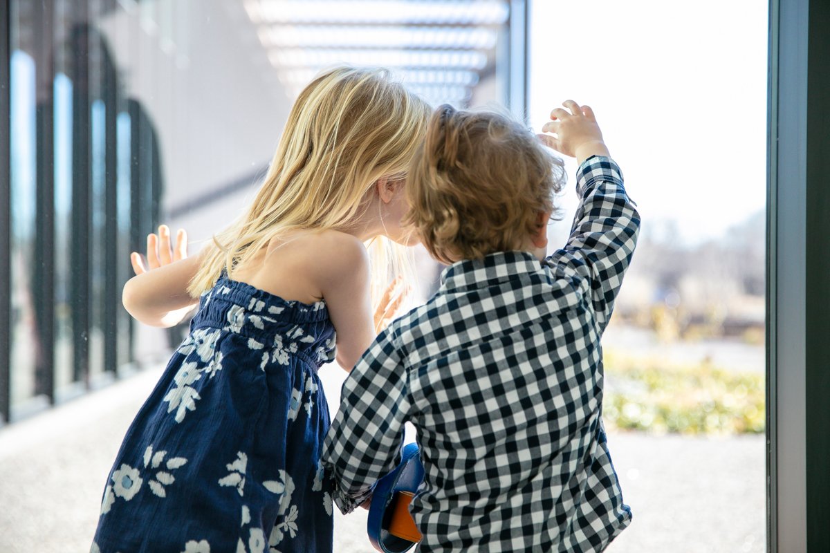 Two children looking out a glass window, one wearing a floral dress and the other in a checkered shirt.