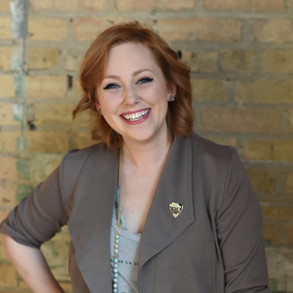 Smiling person with red hair and nose ring wearing a blazer, posing against a brick wall.