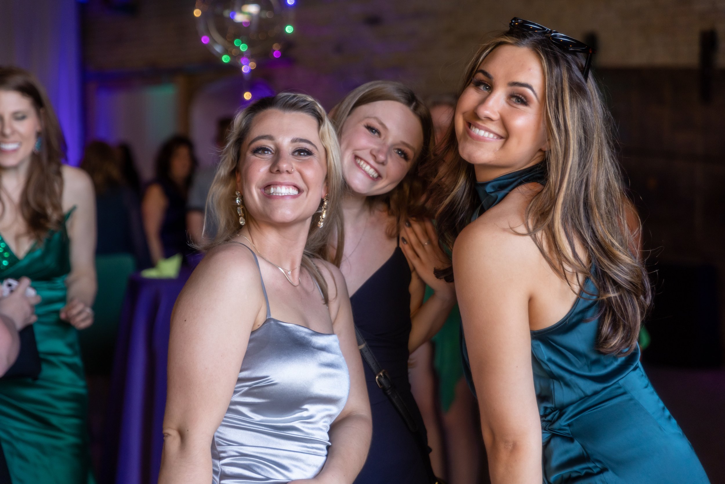 Three women smiling at a party, wearing elegant dresses, with colorful lighting and blurred background.
