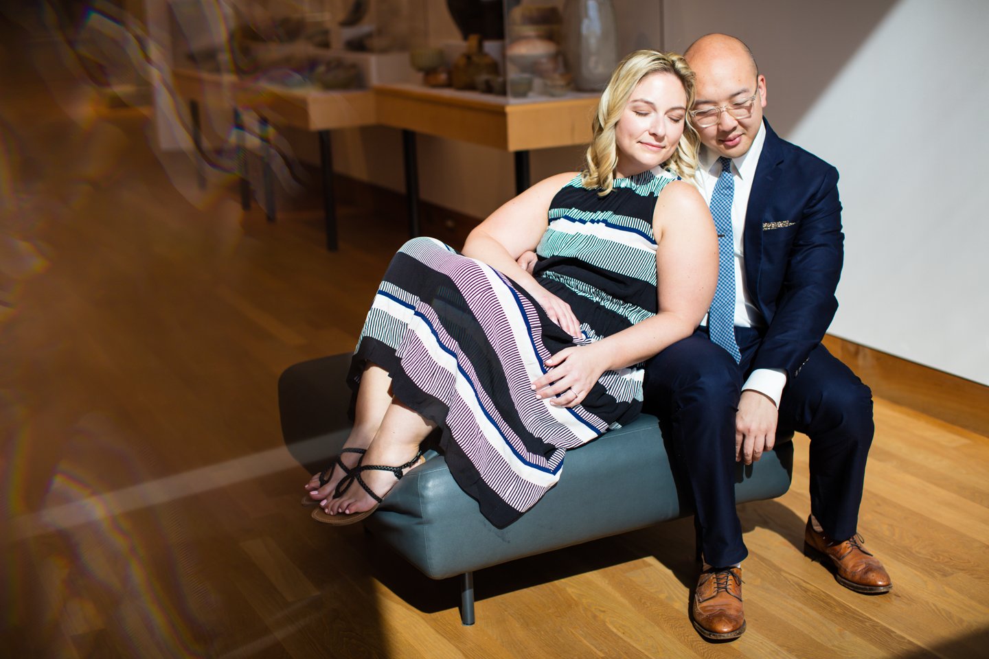 A couple sitting closely on a modern bench in an art gallery, the woman wearing a striped dress and sandals, while the man is dressed in a suit and tie. They appear relaxed and content.